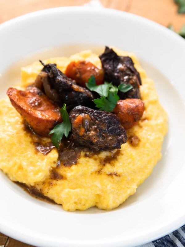White plate filled with yellow polenta and topped with Beef Bourguignon along with carrots and topped with parsley