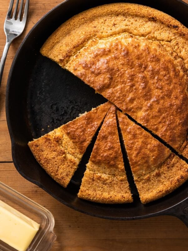 Top down view of slices of cornbread sitting in black cast iron skillet with pat of butter on the side all on wood surface