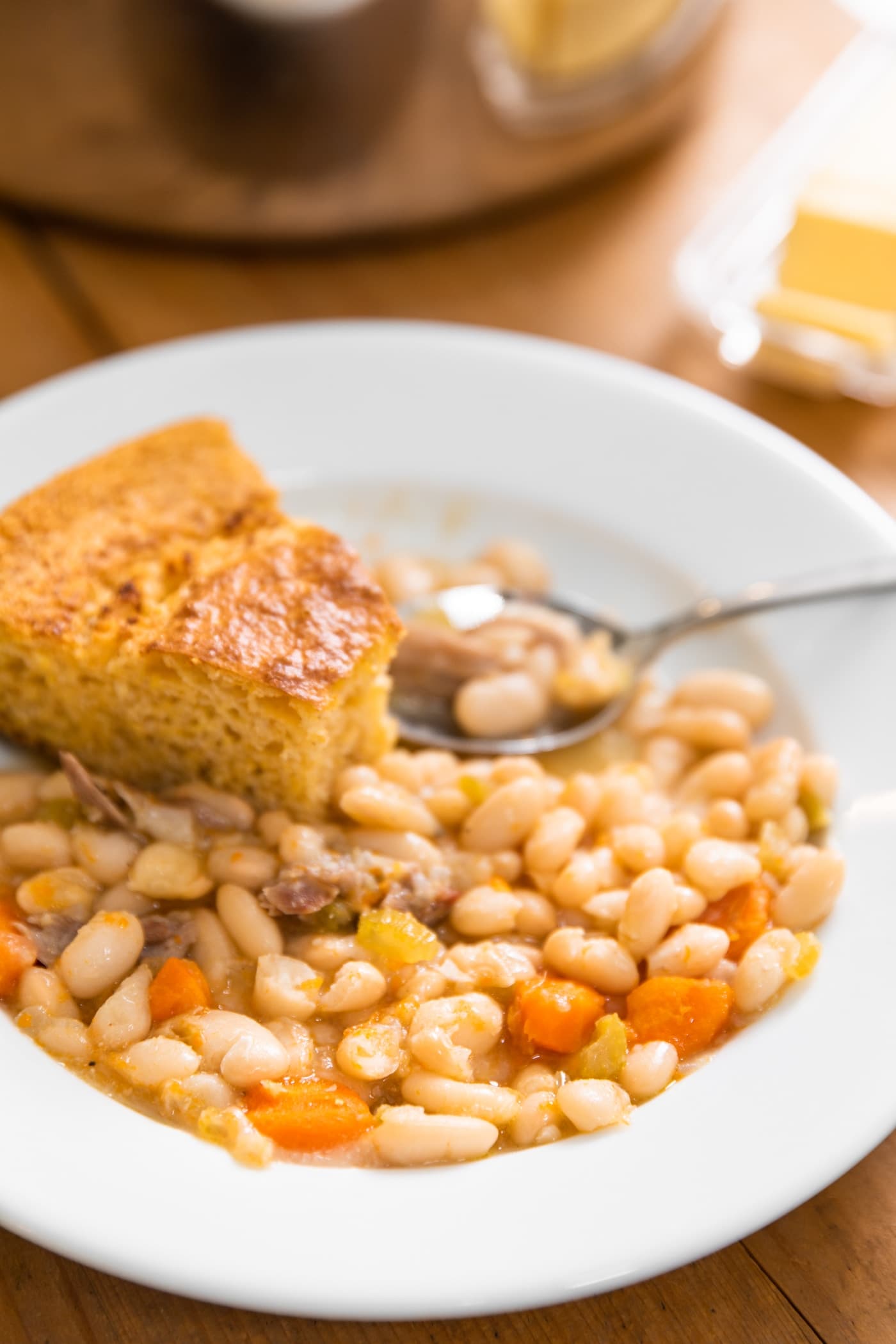 White bowl holding yellow colored bean soup with pieces of ham and carrots with slice of cornbread