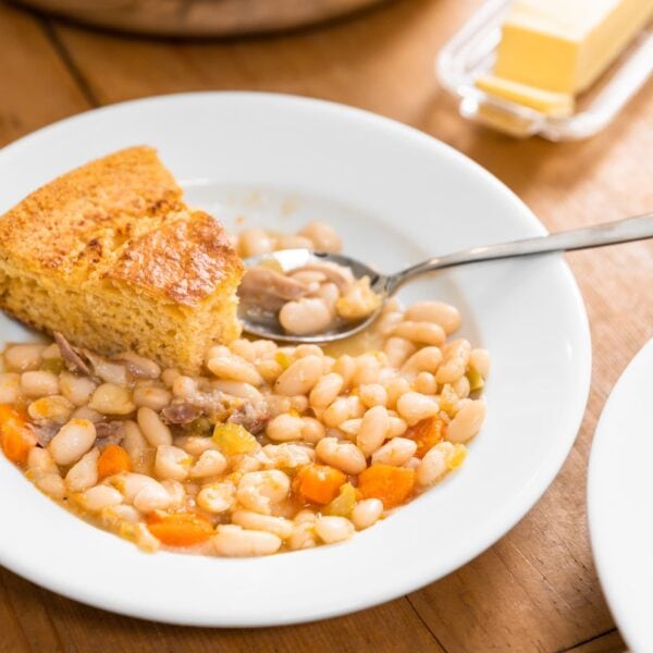 White plate filled with white beans, ham, and carrots with piece of cornbread in amongst soup all on wood board