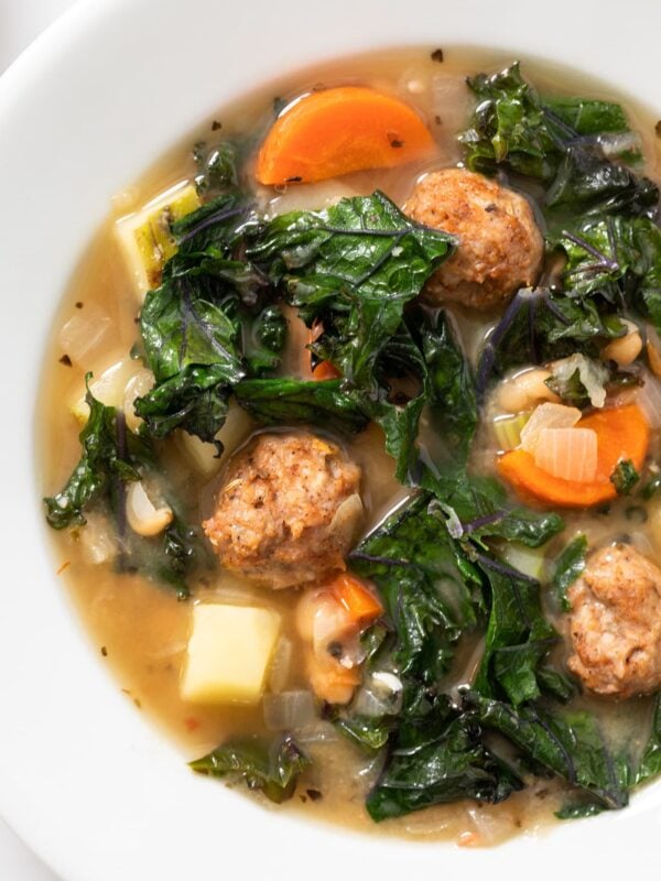 Top down view of white bowl holding light brown soup filled with kale and carrots and meatballs
