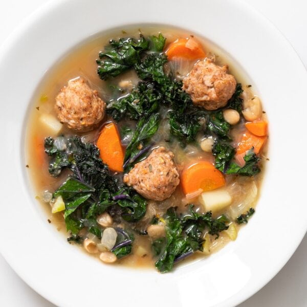 Top down view of white bowl holding light brown soup filled with kale and carrots and meatballs