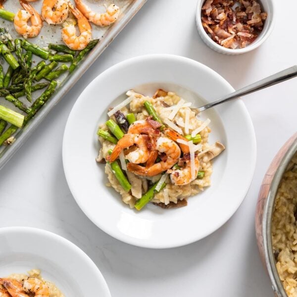 Top down view of white bowl filled with creamy light brown colored risotto with sheet pan filled with asparagus and shrimp to the side