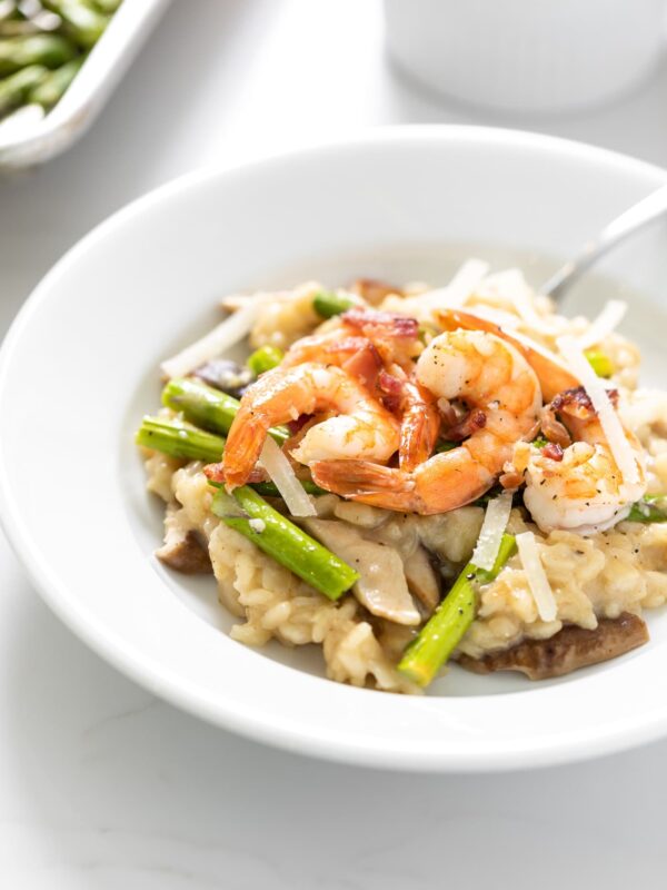 White bowl filled with Parmesan risotto and topped with shrimp and asparagus with sheet pan in background all on white surface