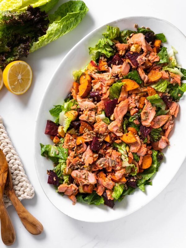 Top down view of white platter filled with green salad topped with pink salmon and roasted vegetables with lemon and extra lettuce to the side all on white counntertop