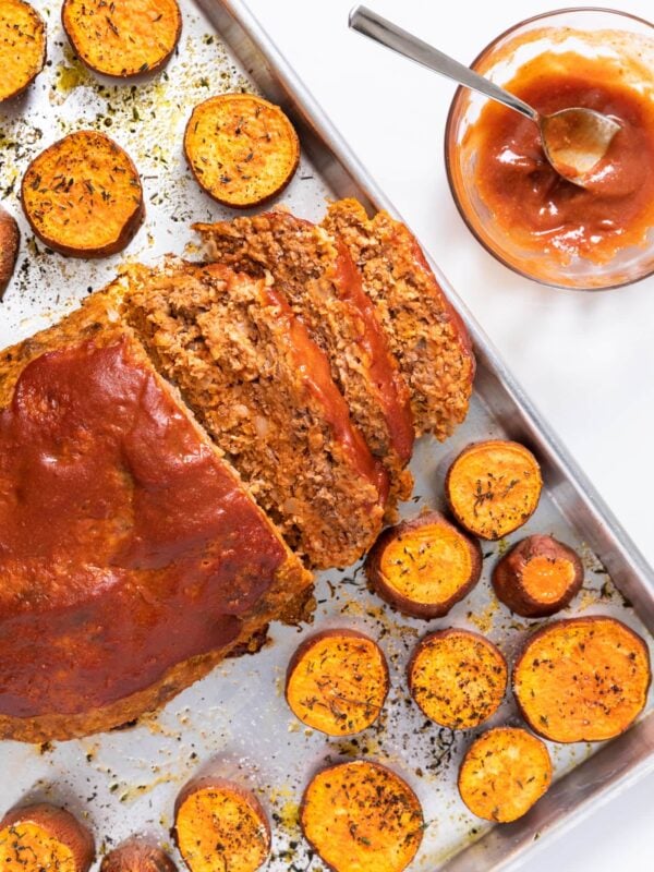 Top down view of white sheet pan filled with meatloaf topped with red ketchup with slices of orange sweet potato all around