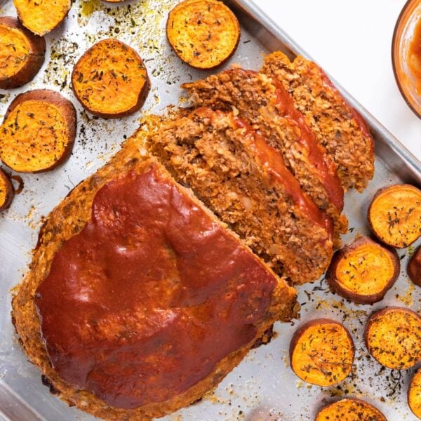 Top down view of meatloaf topped with red ketchup sitting on sheet pan with slices of orange sweet potato freshly baked from the oven
