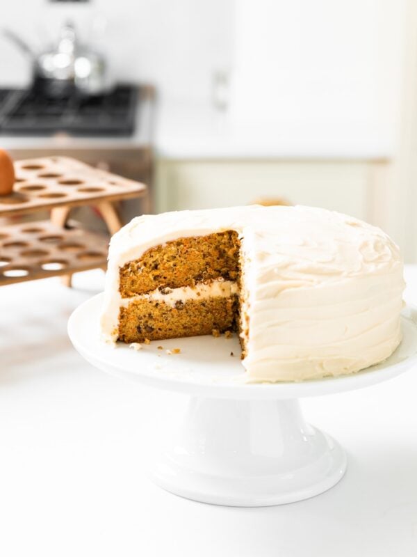 White cake plate with carrot cake with slices missing all sitting on white countertop with egg rack in background