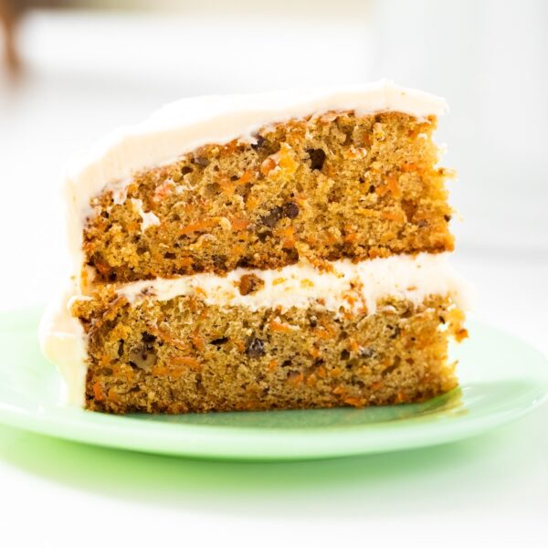 Slice of layered carrot cake sitting on green plate on a white countertop with a white cake plate in background