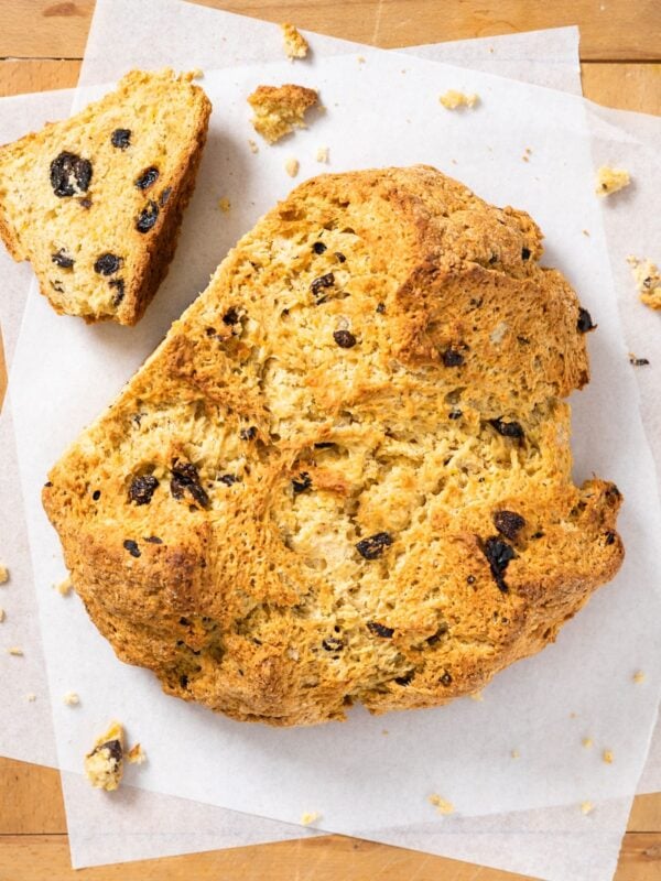 Top down view of circular brown colored loaf of Irish soda bread sitting on white parchment paper with one slice sliced off and laying to the side
