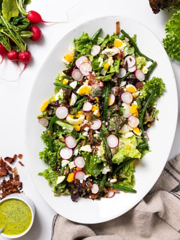 Top down view of white platter filled with green lettuce topped with spring vegetables with extra dressing on the side all on white countertops