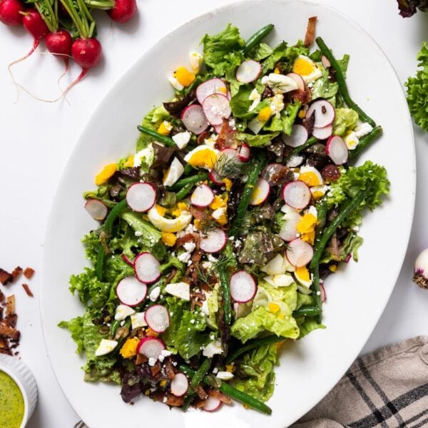 Top down view of white serving platter filled with green butter lettuce topped with fresh spring vegetables and hardboiled egg with extra ingredients sitting all around on white countertop