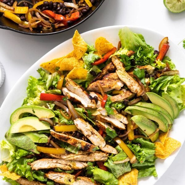 Top down view of white platter filled with salad greens and topped with grilled chicken pieces and grilled fajita vegetables all on white countertop