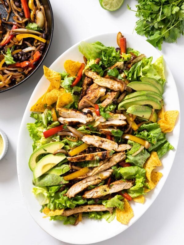 Top down view of white platter filled with salad greens and topped with grilled chicken and tortilla chips with extra grilled vegetables on the side in black skillet