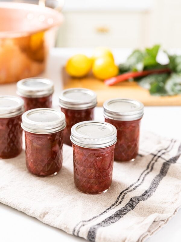 Glass jam jars with lids and rings on top filled with red colored rhubarb jam sitting on cooling cloth with copper colored jam pan in background