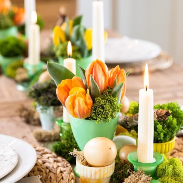 Green vase filled with orange tulips all on wood table with candles lit and white plates set around