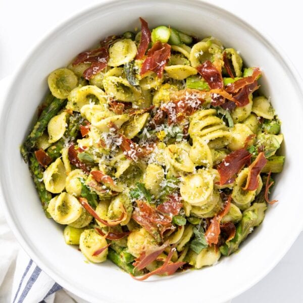 Top down view of yellow pasta covered in pesto sauce and topped with pieces of crispy prosciutto all on white countertop