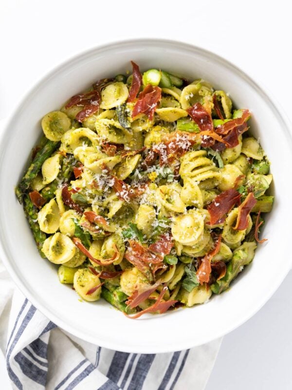 Top down view of large white bowl filled with pesto covered pasta mixed with crispy pieces of prosciutto and asparagus all on white countertop