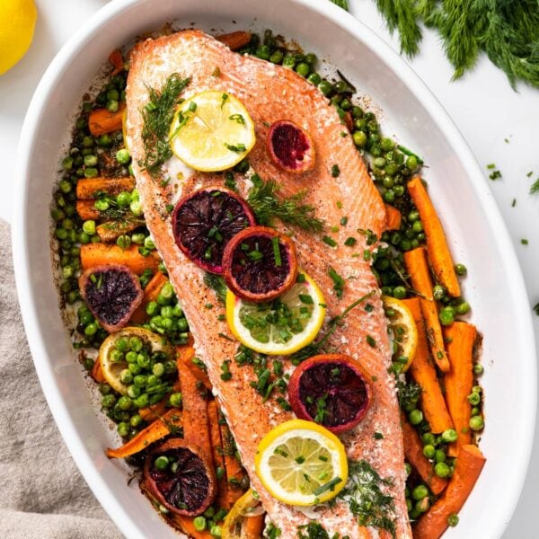 Top down view of long piece of pink salmon sitting on a bed of carrots and peas and topped with slices or orange and lemon with extra dill to the side