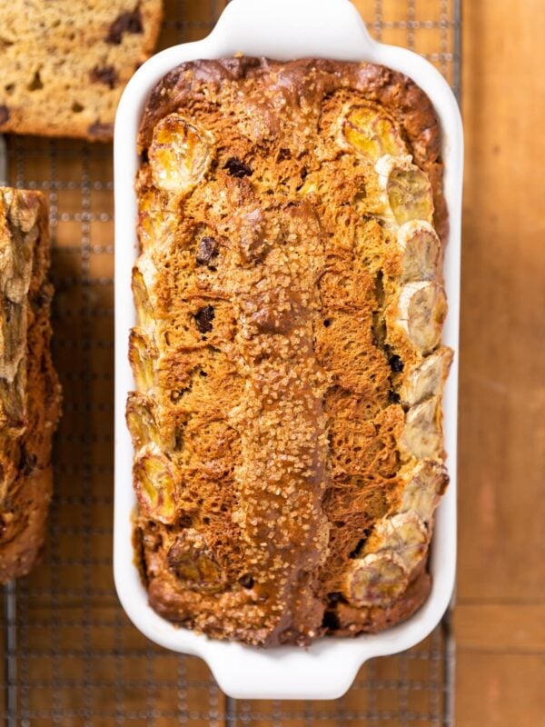 Top down view of ultimate banana bread loaf topped with browned pieces of banana and turbinado sugar in white loaf pan sitting on cooling rack