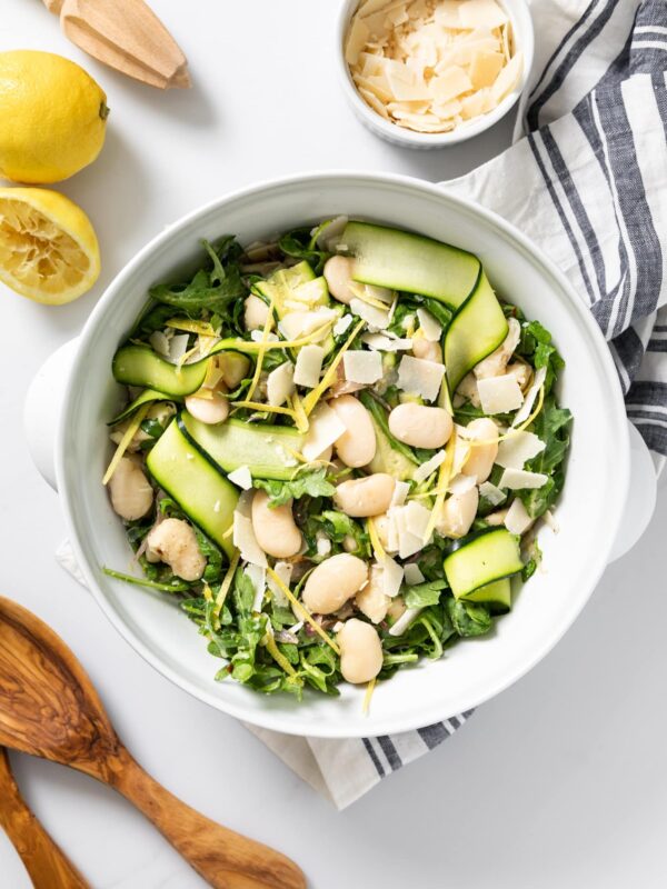 Top down view of white bowl filled with greens and white beans with wooden serving spoons and lemon to the side