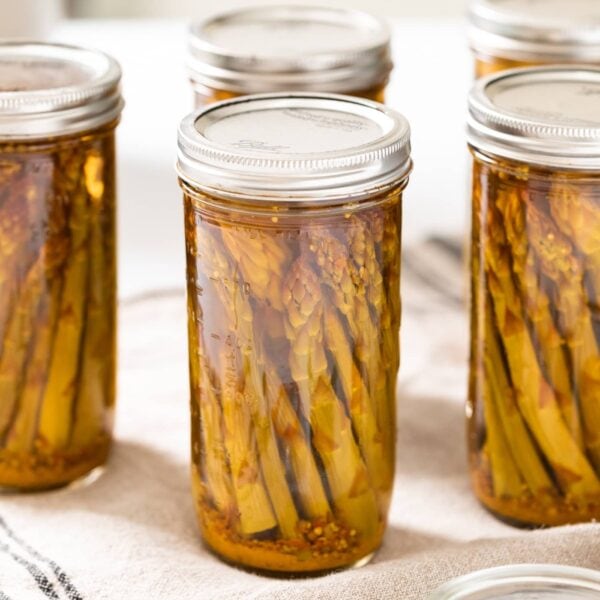 Large glass jars filled with spears of pickled asparagus sitting on cloth on white countertop