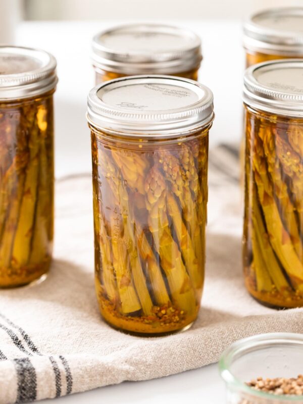 Glass canning jars filled with spears of asparagus all sitting on cloth with open jars of spices around