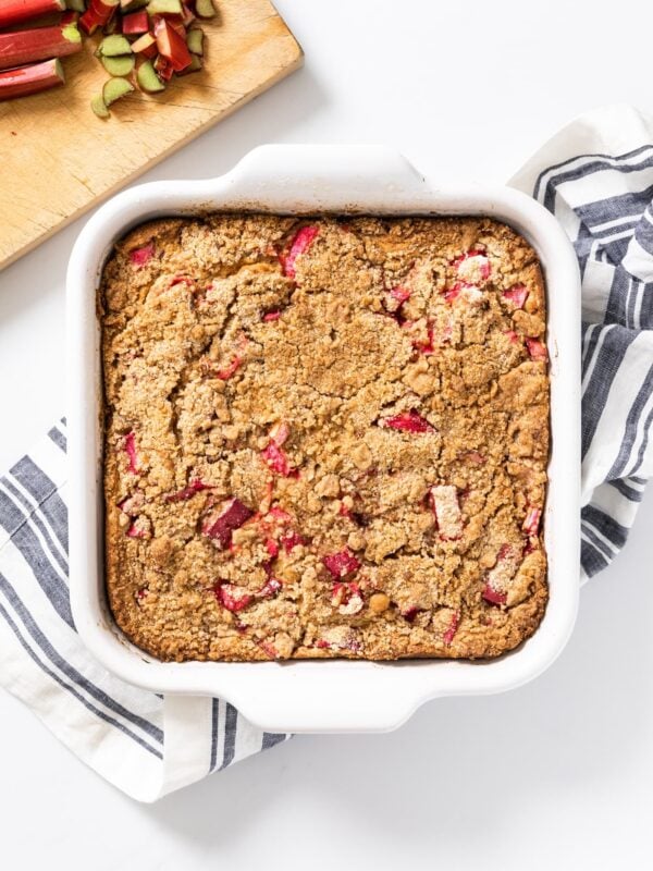 White square baking dish sitting on white surface filled with rhubarb coffee cake with cutting board with sliced rhubarb to the side.