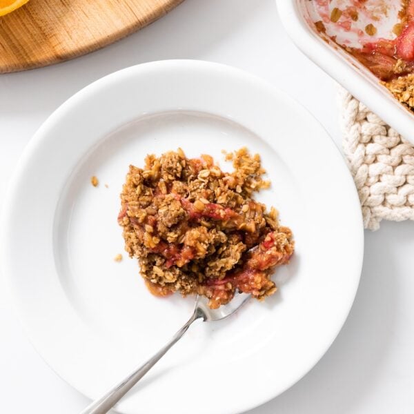 Top down view of white plate filled with brown and red colored rhubarb crisp with baking container sitting beside all on white countertop