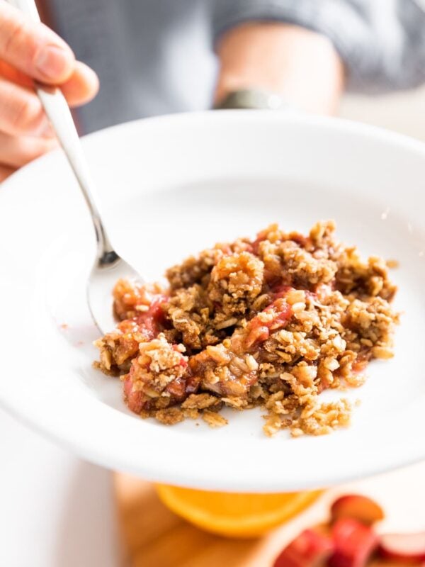 Close up view of red and brown colored rhubarb crisp sitting on white plate with hand holding fork