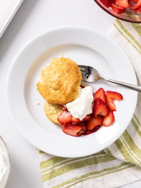 Top down view of white plate filled with strawberry shortcake and biscuit and topped with whipped cream with green napkin underneath