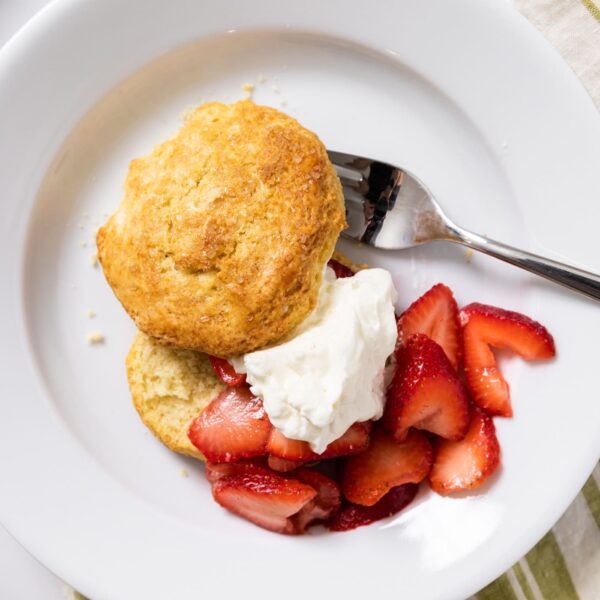Top down view of white plate with biscuit, strawberries, and whipped cream with striped green napkin underneath