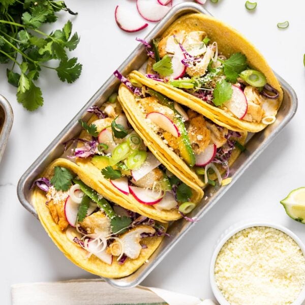 Top down view of metal container holding four corn tortillas filled with cod and topped with radish and coleslaw