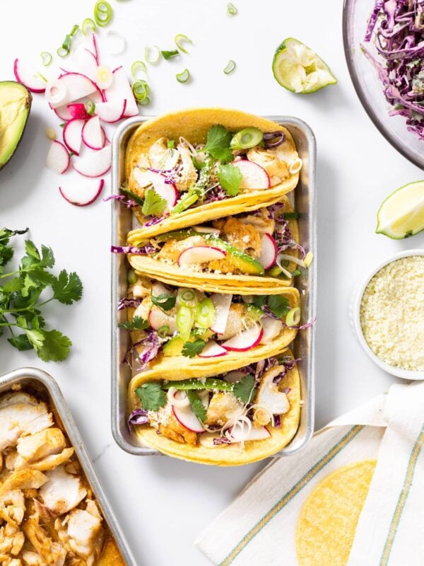 Top down view of four tacos sitting in metal container with toppings all around including tortillas and purple coleslaw