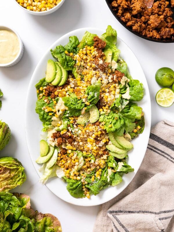 Top down view of white platter sitting on white countertop filled with lettuce and topped with corn and chorizo with extra toppings sitting all around