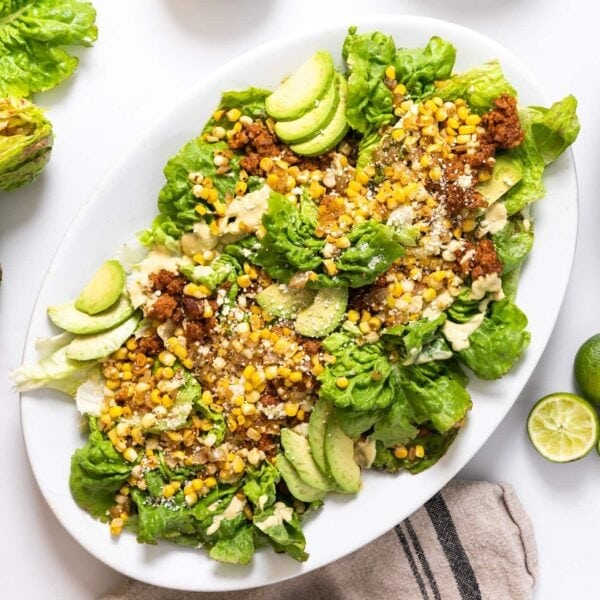 Top down view of white platter sitting on white countertop filled with pieces of lettuce and topped with avocado and sweet corn and chorizo with extra toppings all around