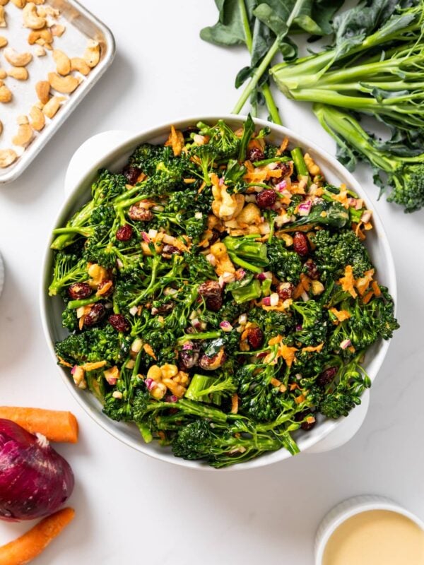 Top down view of white bowl holding broccoli florets topped with cashews with extra vegetables sitting around along with peach colored dressing