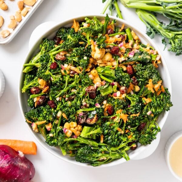Top down view of white bowl filled with florets of broccoli tossed in dressing with dried cranberries and cashews on top with other ingredients sitting around on white countertop