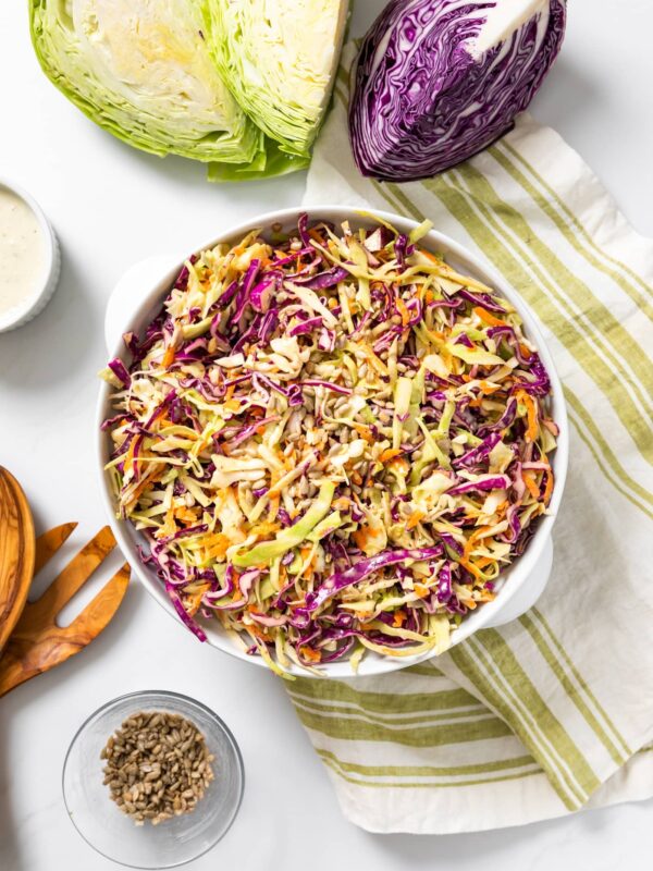 Top down view of white bowl sitting on white countertop filled with brightly colored coleslaw in both green and purple with extra cabbage around