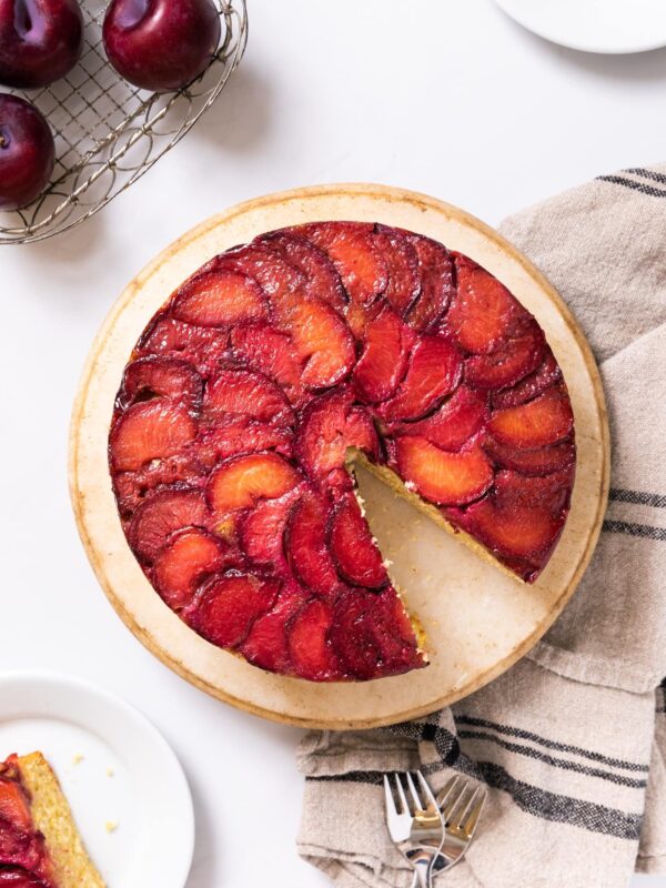 Top down view of bright red colored plum cake sitting on cake platter on white countertop with extra piece of cake and plums