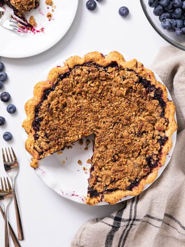 Top down view of circular blueberry pie sitting on white countertop with slice taken out and sitting on white plate with extra bowl of blueberries