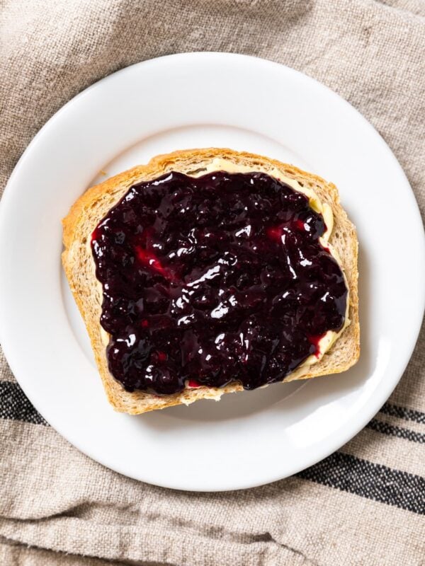 Top down view of slice of bread with butter and blueberry jam spread on top sitting on white plate on towel
