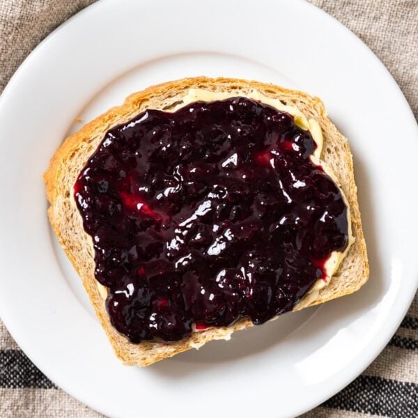 Top down view of slice of brown bread sitting on white plate with butter and blueberry jam spread on top
