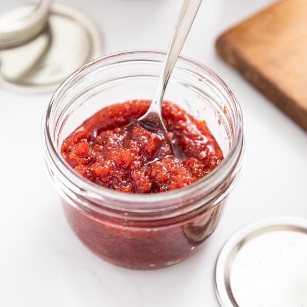 Small glass jar sitting on white countertop filled with red colored cherry jam with lids and rings all around and wood cutting board in background