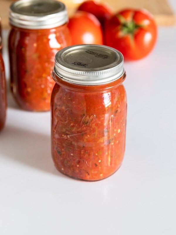 Glass canning jar filled with red colored salsa with lid and ring on top sitting on white countertop with tomatoes in background