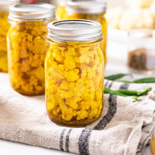 Glass canning jar filled with yellow colored pickled curry cauliflower sitting on towel with other glass jars in background with peppers and spices all around