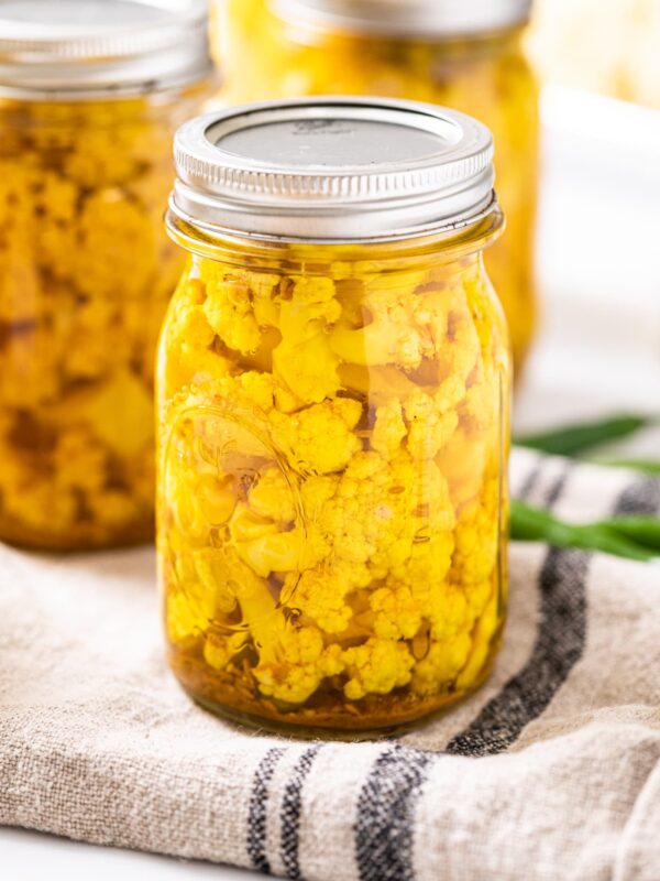 Glass jar with lid and ring on top filled with yellow cauliflower sitting on towel with other filled jars in background