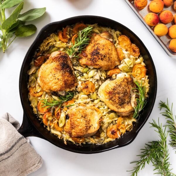 Top down view of black skillet with cooked and browned chicken thighs sitting amongst orzo and apricots and herbs with extra apricots and herbs spread around
