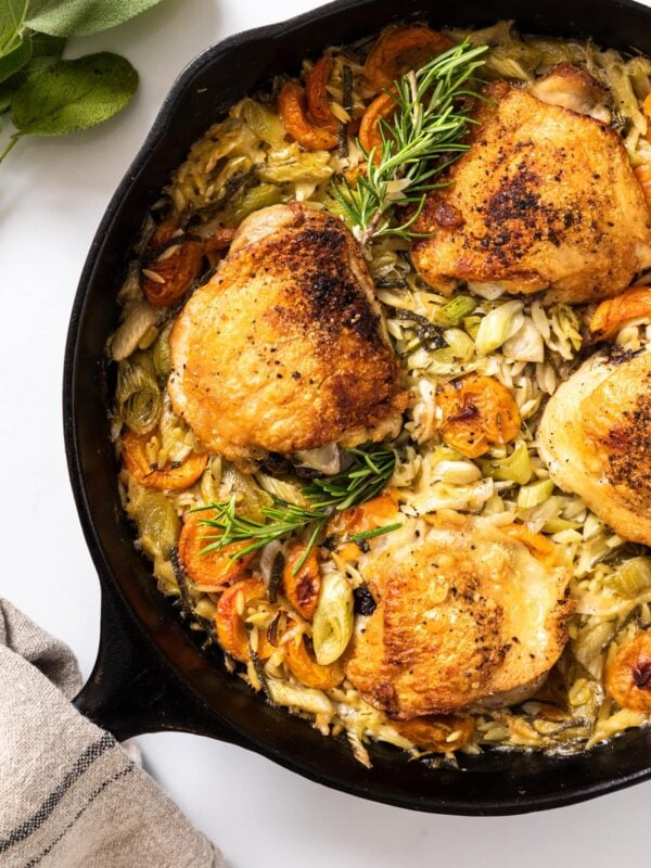 Top down view of black skillet sitting on white countertop with cooked chicken thighs and orzo with apricots and herbs