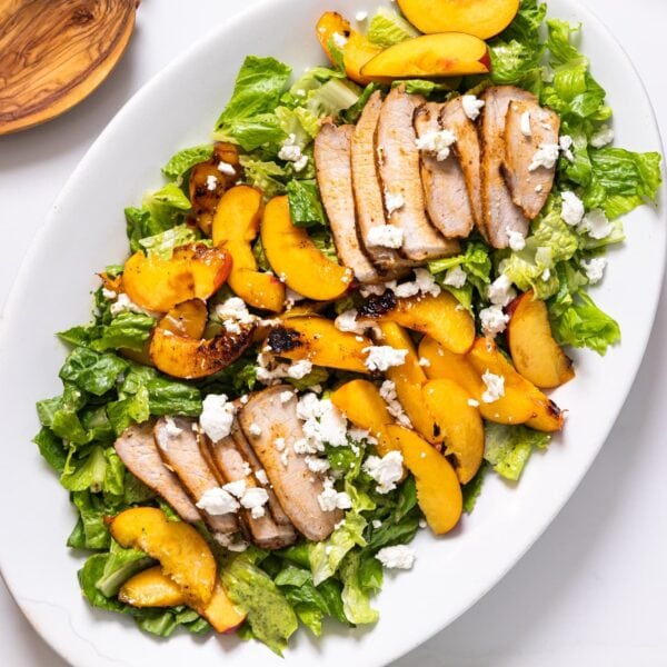 Top down view of white platter filled with lettuce and slices of nectarines and pork topped with goat cheese all on white countertop with components of salad all around